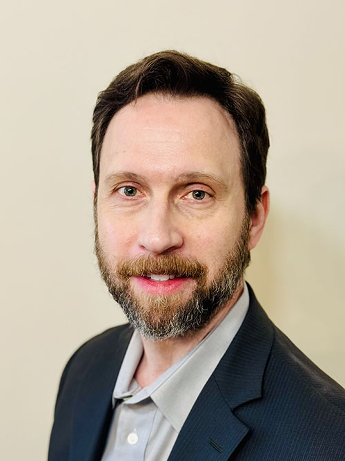 The image shows a man wearing glasses, a blue shirt, and a white coat standing in front of a door with a sign that reads Dental Office.
