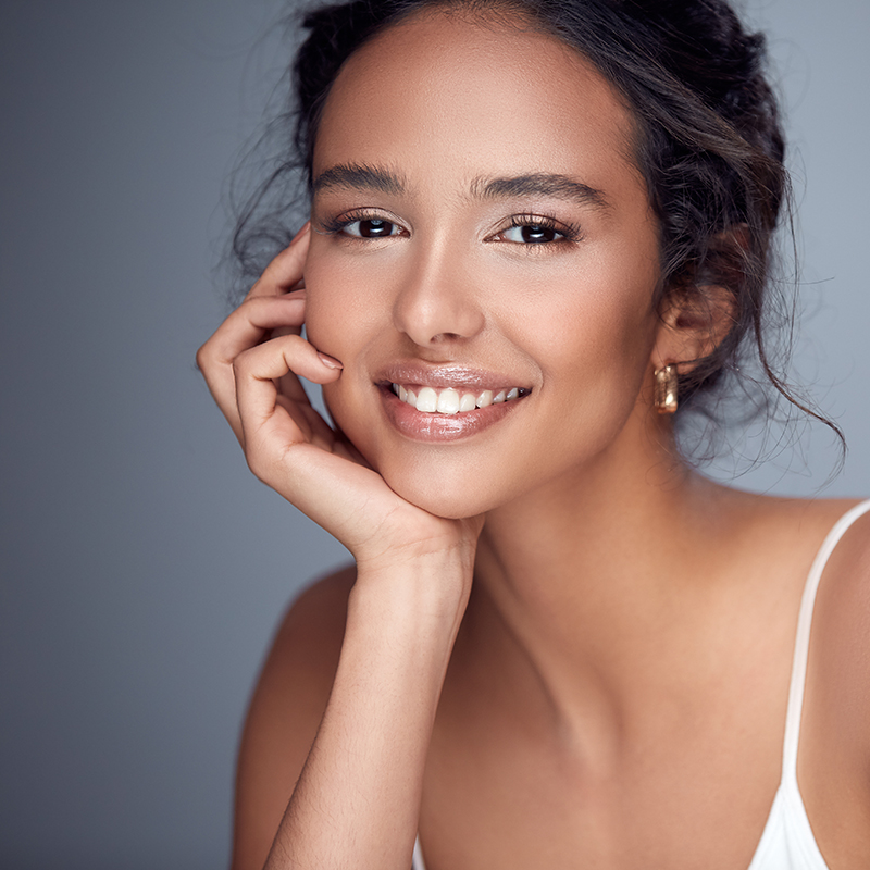 The image features a young woman with a radiant smile, posing for the camera. She has light skin and is wearing what appears to be a white top. Her hair is styled in an updo, and she has her left hand placed on her chin. The background of the photo is neutral, providing a clear focus on her.
