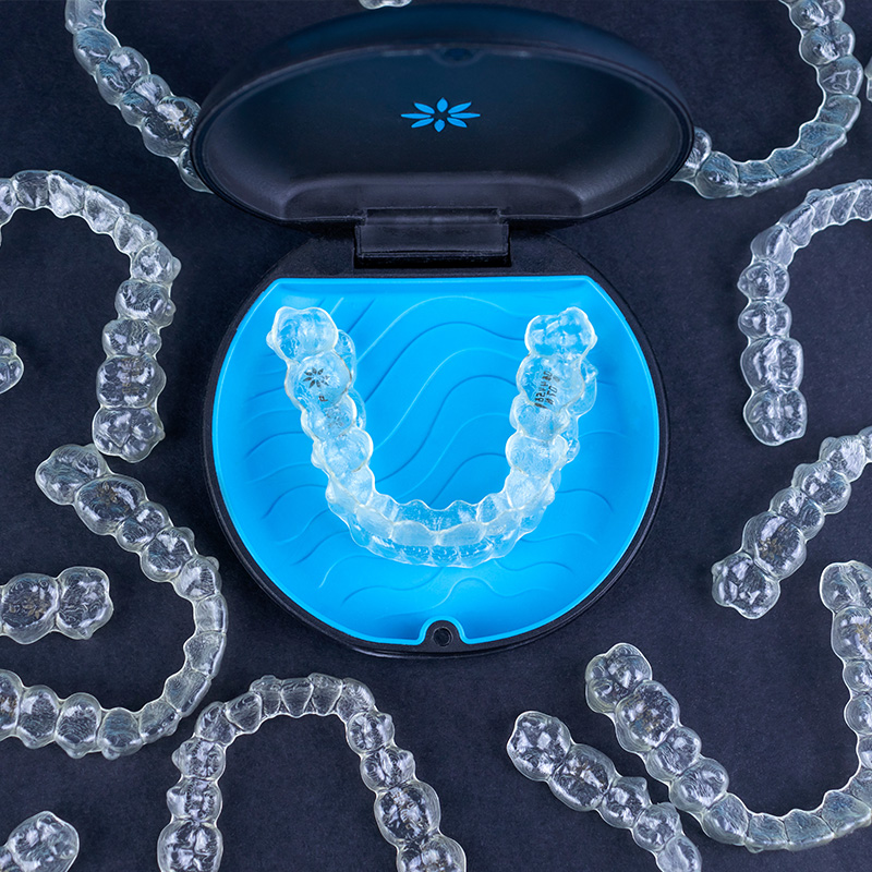 An image of a transparent plastic tray containing multiple clear orthodontic aligners, with a blue smile-shaped tray and a box of additional trays in the background.