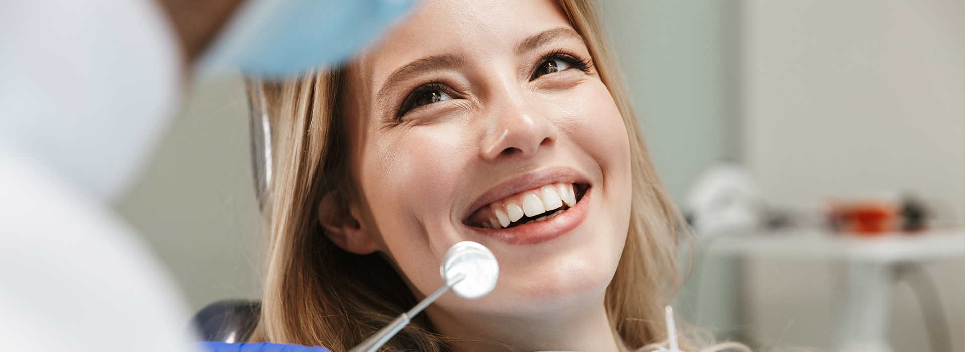 The image shows a woman in a dental office, smiling at the camera, while a dentist is examining her teeth.