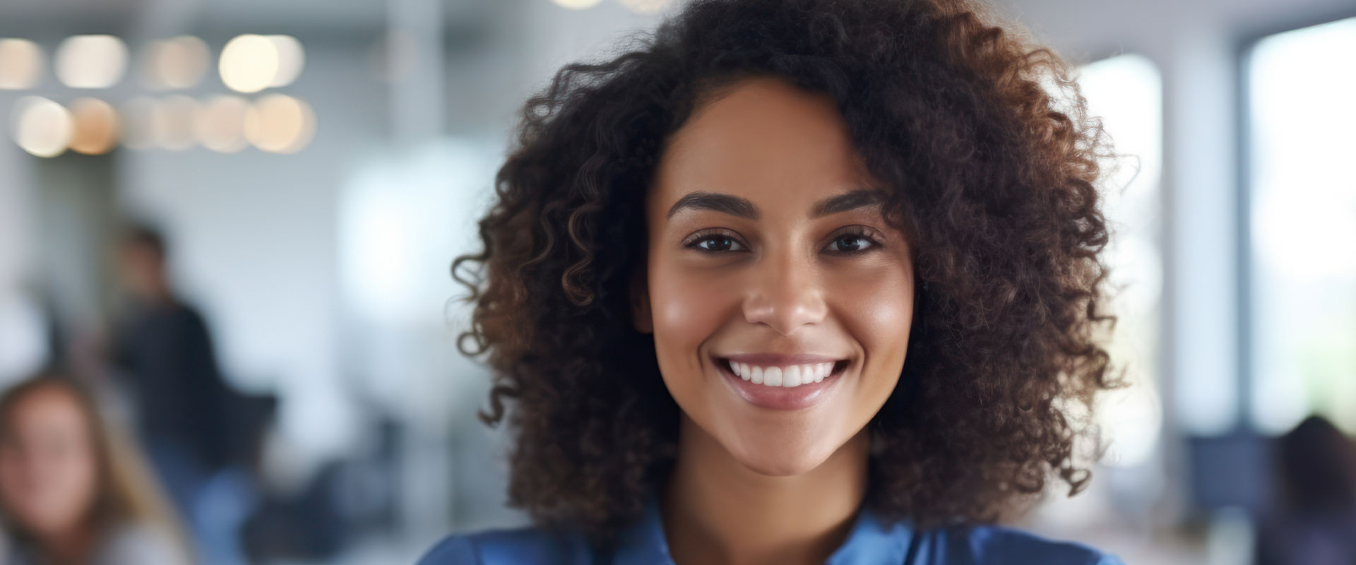 The image shows a smiling woman with curly hair, wearing a dark top, standing in an office environment.