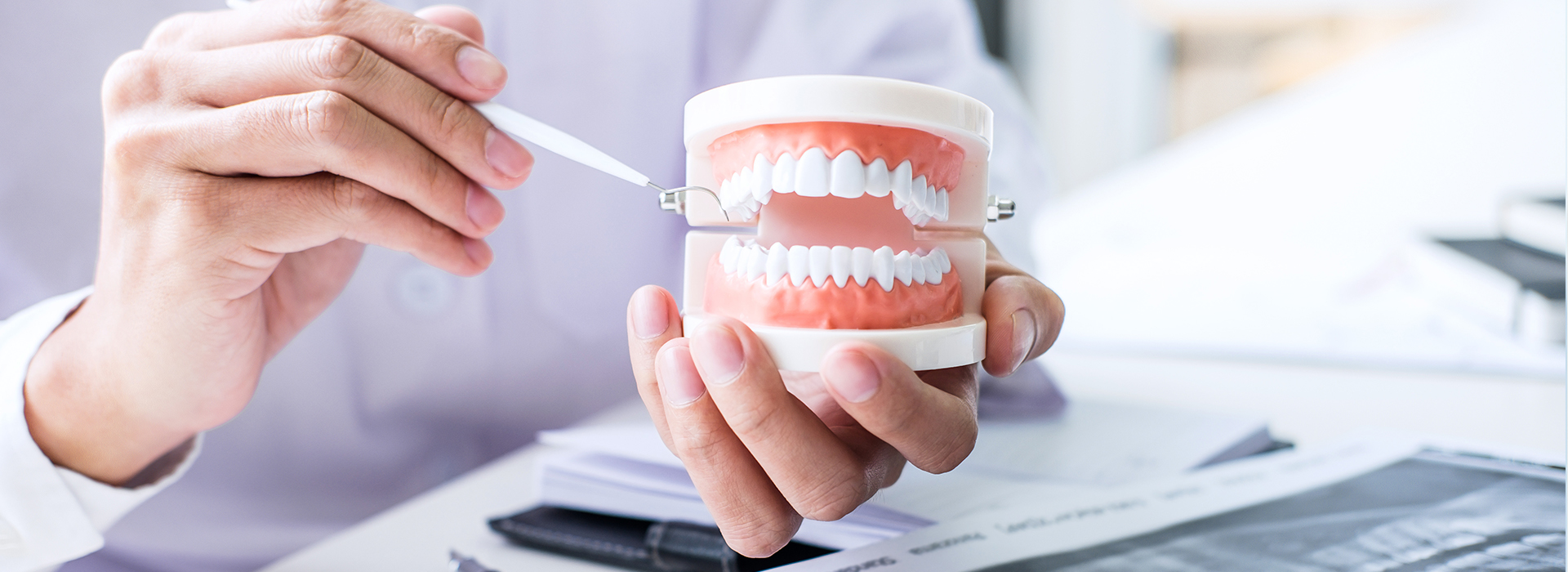 The image shows a person s hand holding a toothbrush with a toothpaste tube and a toothbrush head, placed in front of a cup that contains a model set of teeth. The background is blurred but includes what appears to be a desk with papers and a computer monitor.
